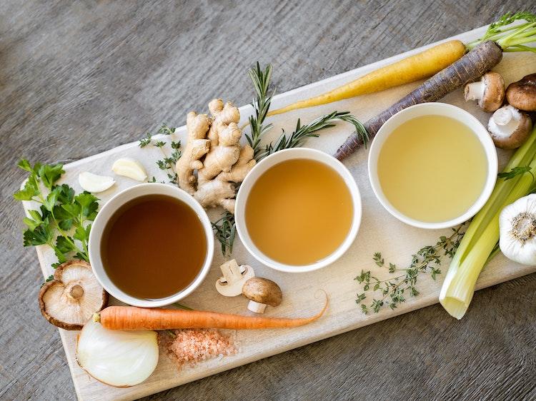 Three different broths on a cutting board with celery, onion, carrots, and mushrooms