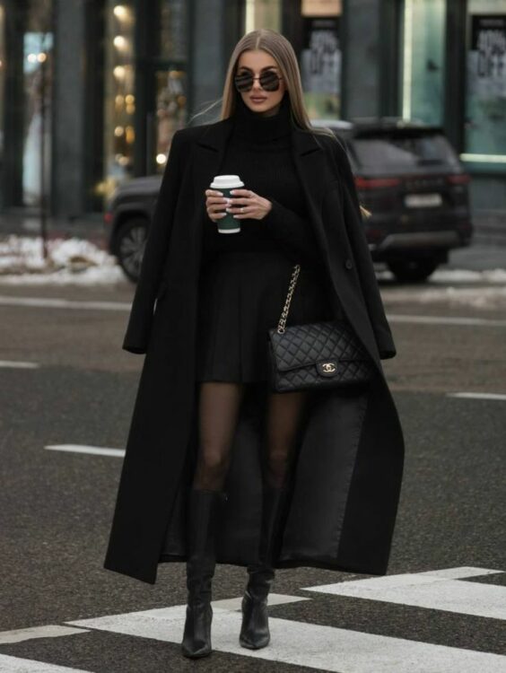 Model wears sunglasses, a black trench coat, and a black dress