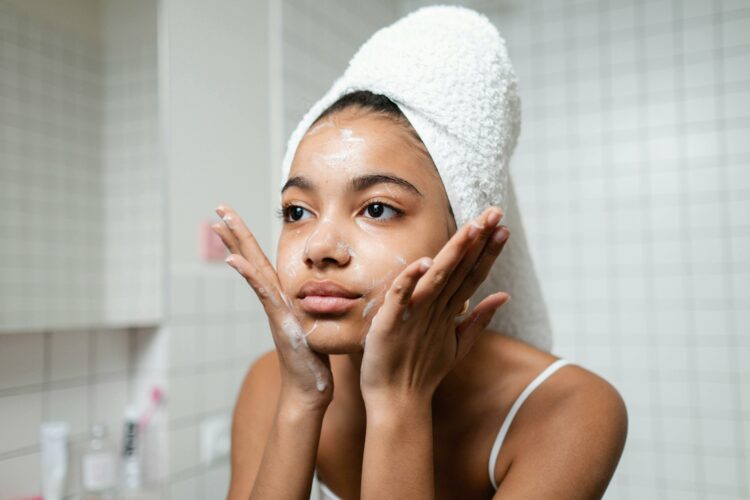 Person with their hair in a towel washes their face in the mirror